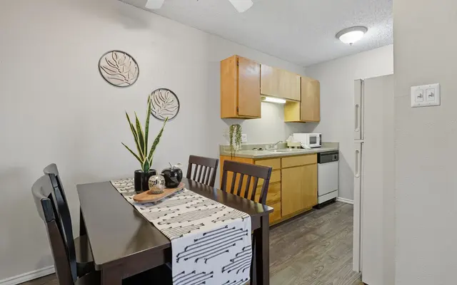 A modern kitchen dining area featuring a wooden table with chairs, a table runner, and a potted plant. The kitchen has wooden cabinetry, appliances, and a window for natural light.