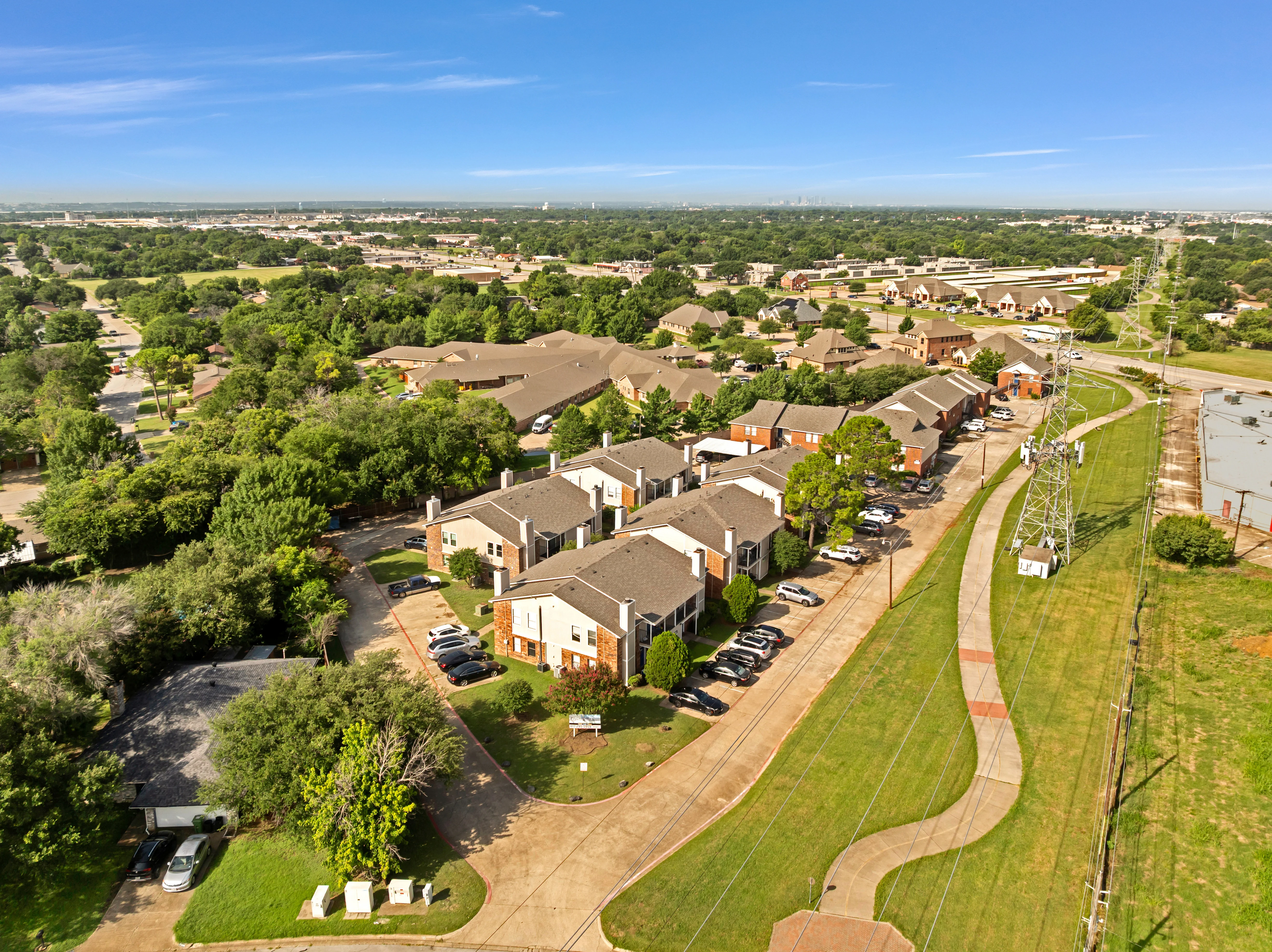 Aerial view of an apartment complex surrounded by trees and green spaces, with a clear blue sky above.