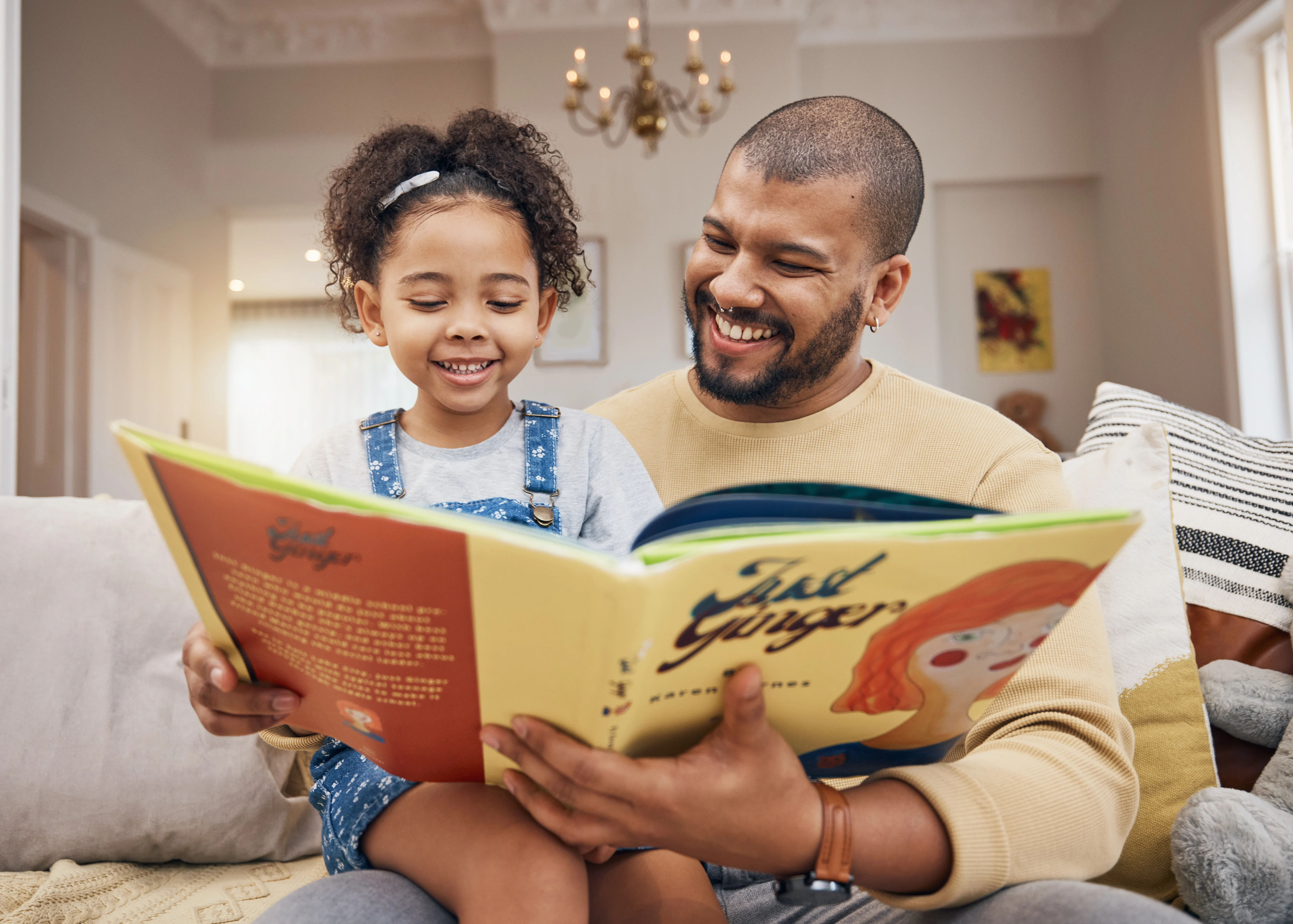 A joyful father and daughter reading a colorful children's book together on a couch. The girl is smiling and pointing at the book, while the father looks at her happily.