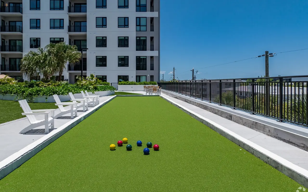 A vibrant lawn bowling area featuring a long, green synthetic grass surface with colorful balls scattered on it. White lounge chairs are positioned nearby, and a modern apartment building is in the background under a clear blue sky.