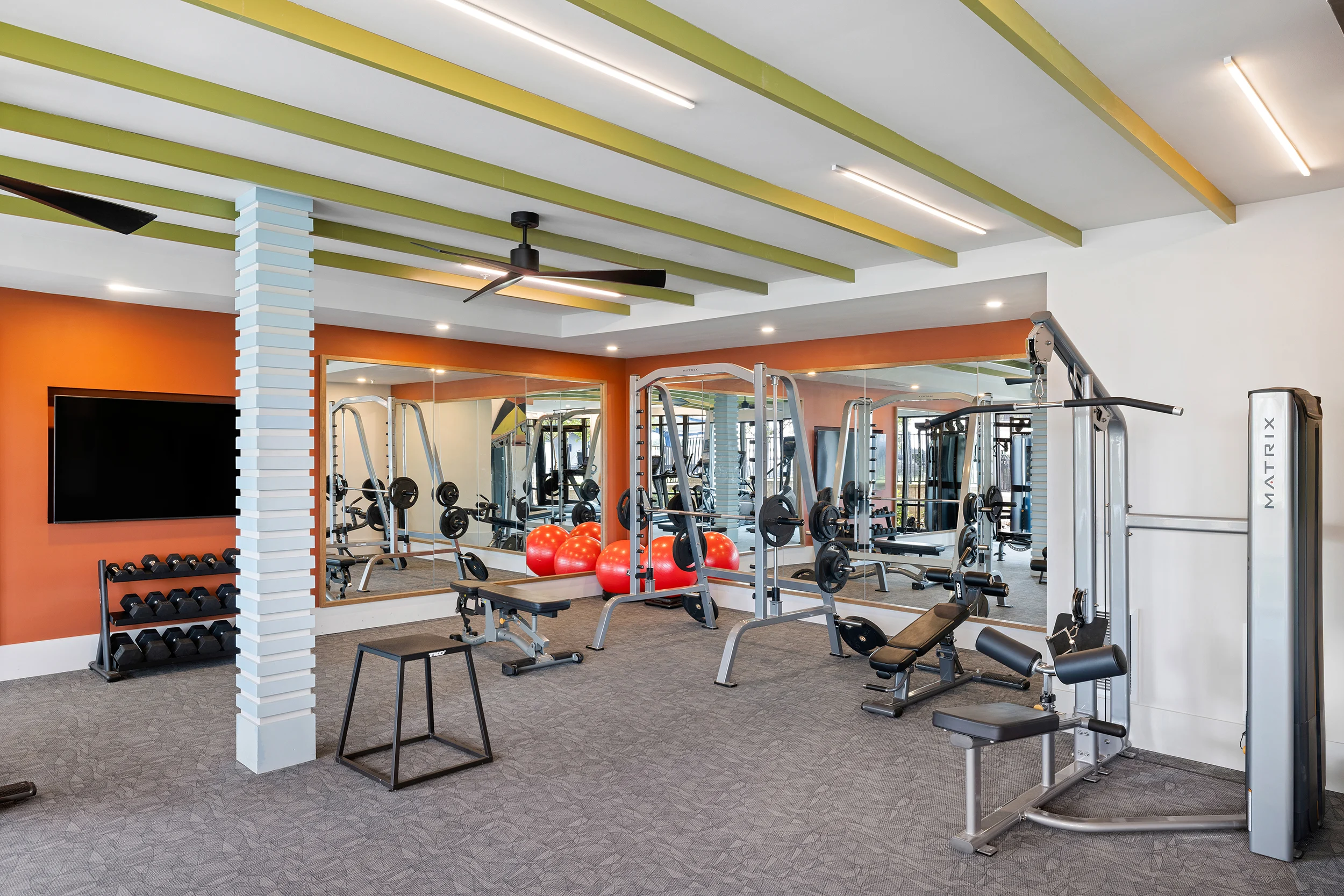 A modern gym interior featuring workout equipment, mirrors, and a television. The gym has a warm color scheme with green ceiling beams and an orange accent wall.