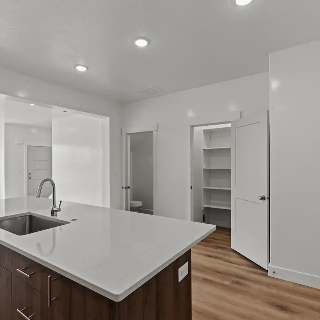 A modern kitchen with a large island, featuring a sink and wooden cabinets, set in a bright room with white walls and light wood flooring.