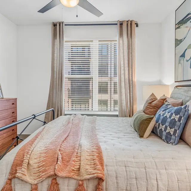 A cozy bedroom featuring a bed with decorative pillows, a blanket draped over the side, a wooden dresser, a mirror, and large windows adorned with light curtains.