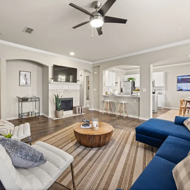 Modern Living Room Design A modern living room featuring a blue sectional sofa, white chairs, a round wooden coffee table, and a light-colored area rug with stripes. In the background, there is a fireplace, a television, and a dining area with a table and chairs.