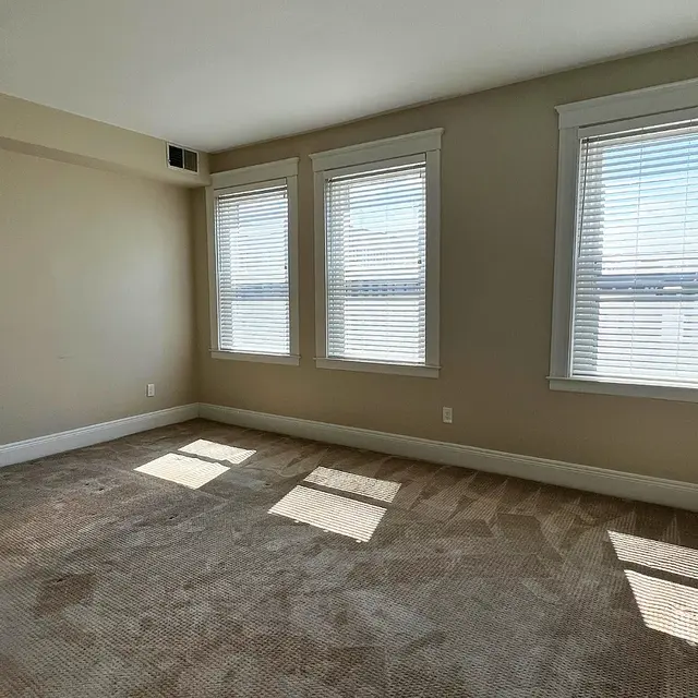 An empty room with beige walls and large windows allowing natural light to enter. The carpet is light brown and the floor is unoccupied, creating a minimalistic atmosphere.