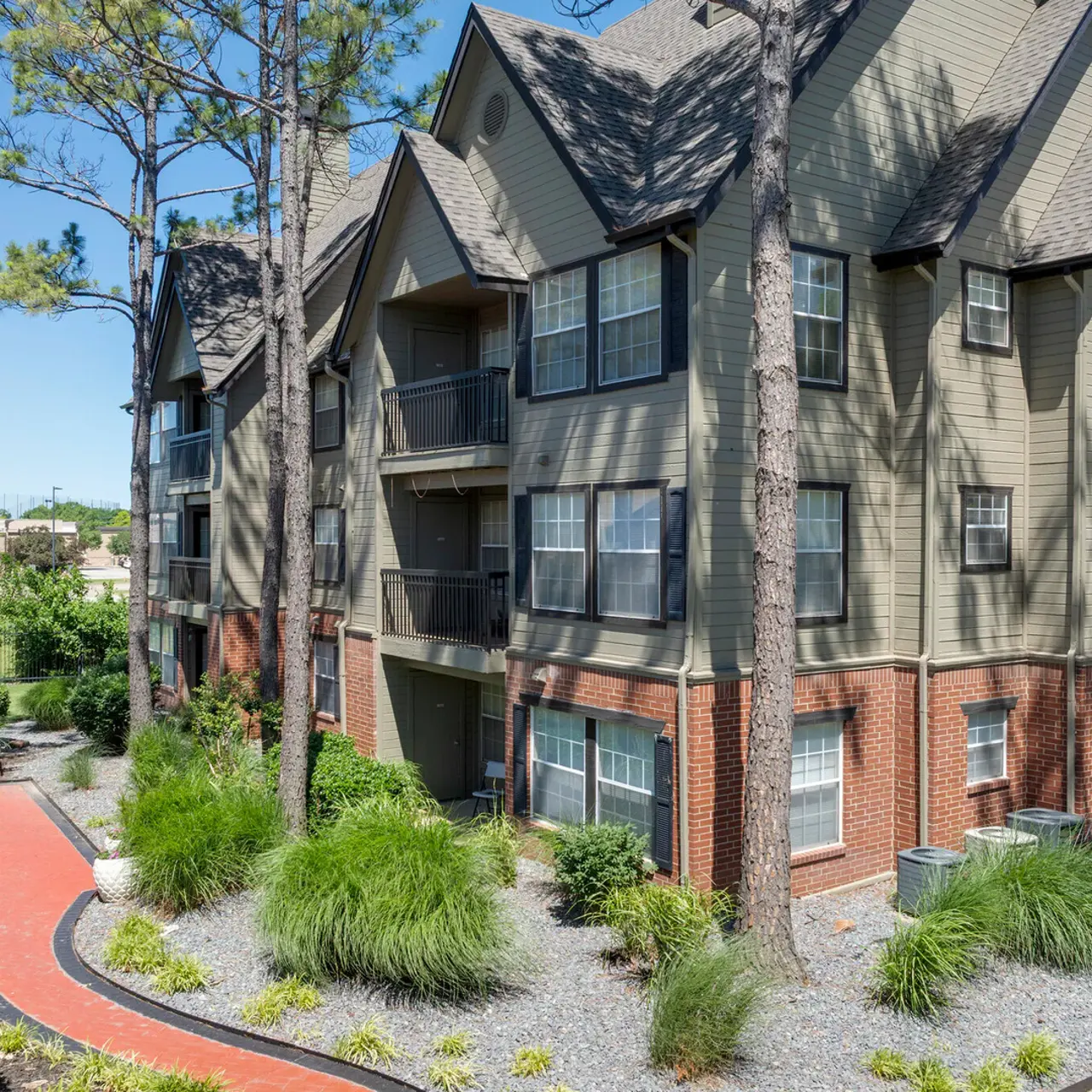 Apartment Complex Exterior View A view of a multi-story apartment complex surrounded by greenery and trees, with a paved walkway and a patio area visible on the left.