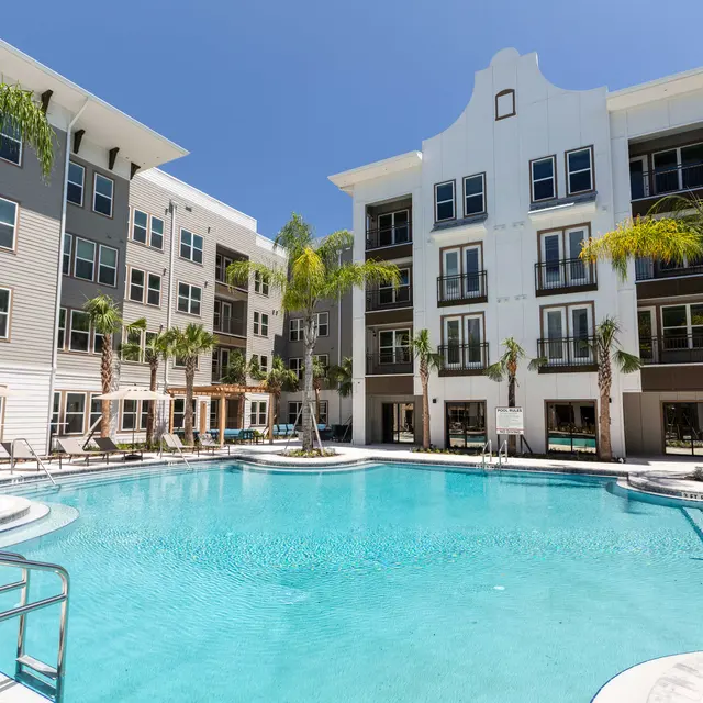 A sunny apartment complex with a swimming pool surrounded by palm trees and lounge chairs.