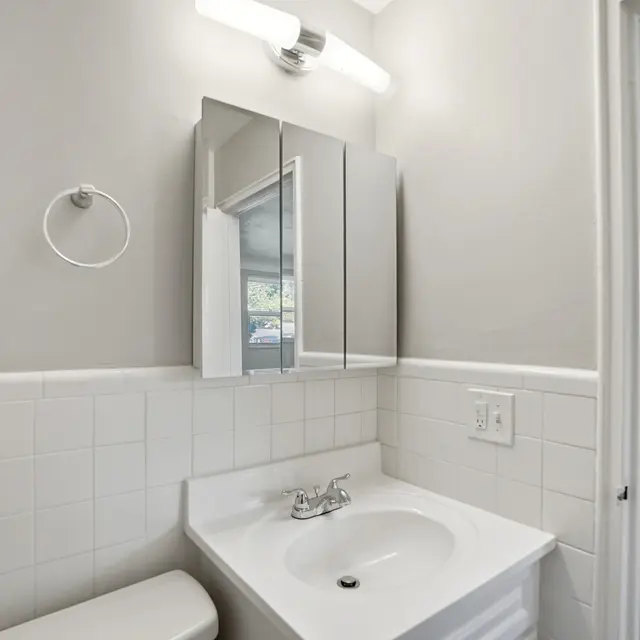 A modern bathroom featuring a white sink with a chrome faucet, a wall-mounted mirror cabinet, and light grey walls. The space is bright with a ceiling light and a tiled backsplash.