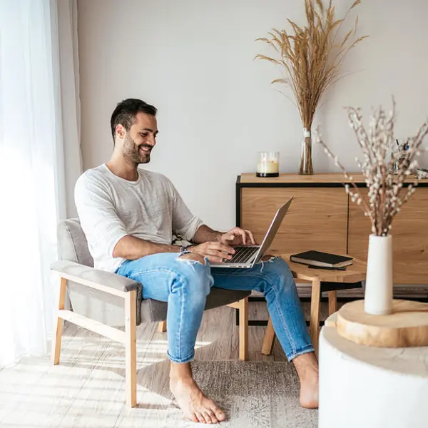A man sits in a stylish room, using a laptop while smiling. Natural light streams in, enhancing the cozy atmosphere.