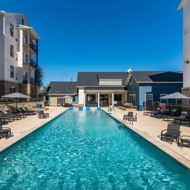 A rectangular swimming pool surrounded by lounge chairs and umbrellas, located in a courtyard of a modern apartment complex.