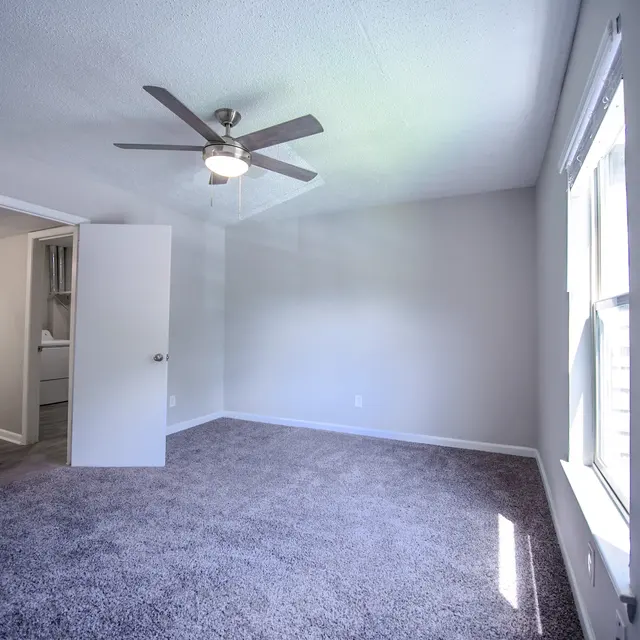 A spacious, empty bedroom with gray carpet, a ceiling fan, and windows letting in natural light. A door leads into another room.
