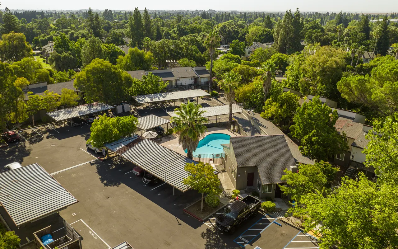 Aerial view of a residential apartment complex surrounded by lush greenery and trees, featuring a central pool area and car parking spaces.
