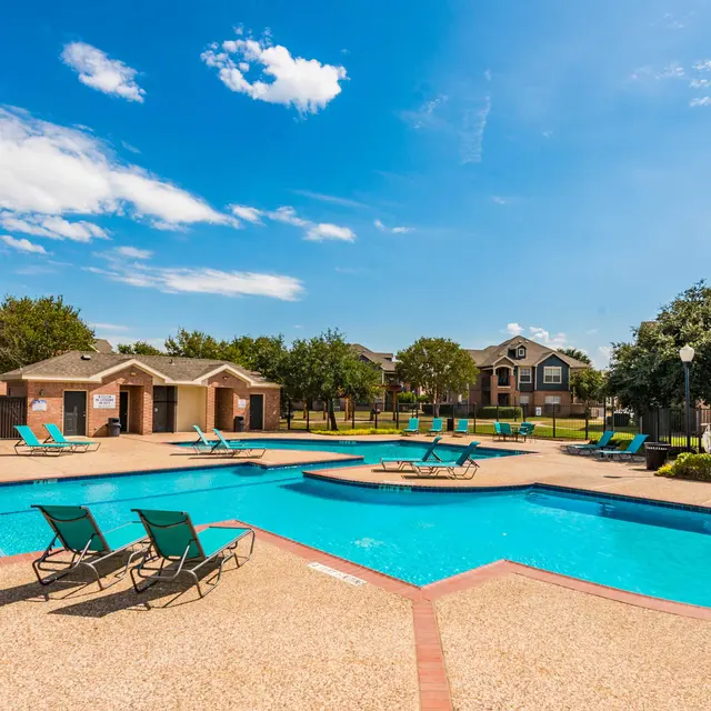 A beautifully landscaped pool area in an apartment complex under a clear blue sky. Lounge chairs are arranged around the pool, with greenery and buildings in the background.