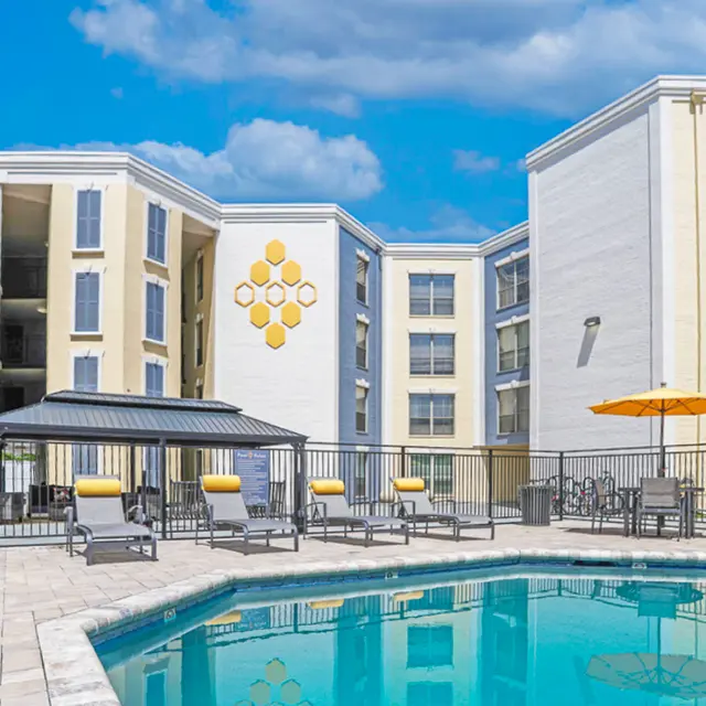 A modern apartment complex featuring a swimming pool surrounded by lounge chairs, with an umbrella and a gazebo. The building has a bright yellow and white exterior with the name 'THE HIVE @ 3RD' displayed prominently.