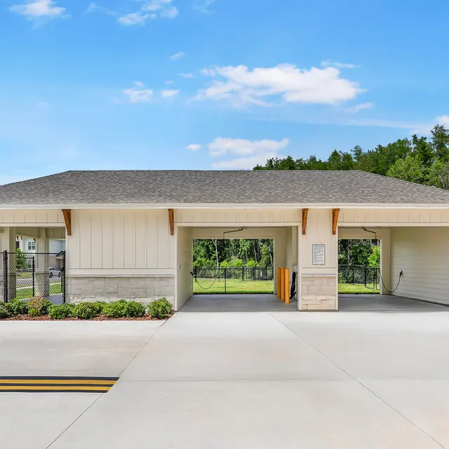 Modern Entry Gate A modern entry gate building with a sloped roof and wooden accents, surrounded by landscaped greenery and trees in the background. A stop sign is visible in front.