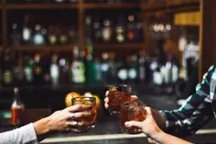 Three people holding their glasses of cocktails up in celebration, fully stocked bar behind them, bottles and glasses.