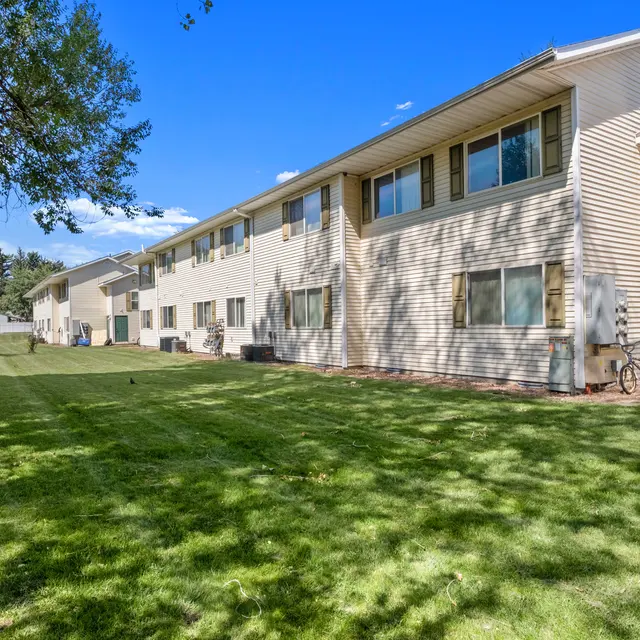 Rear view of Courtyards at Sheridan apartments building with a large, well-maintained grassy backyard area, ideal for outdoor activities. The building has multiple windows and shaded areas provided by nearby trees, creating a serene and open environment for residents.