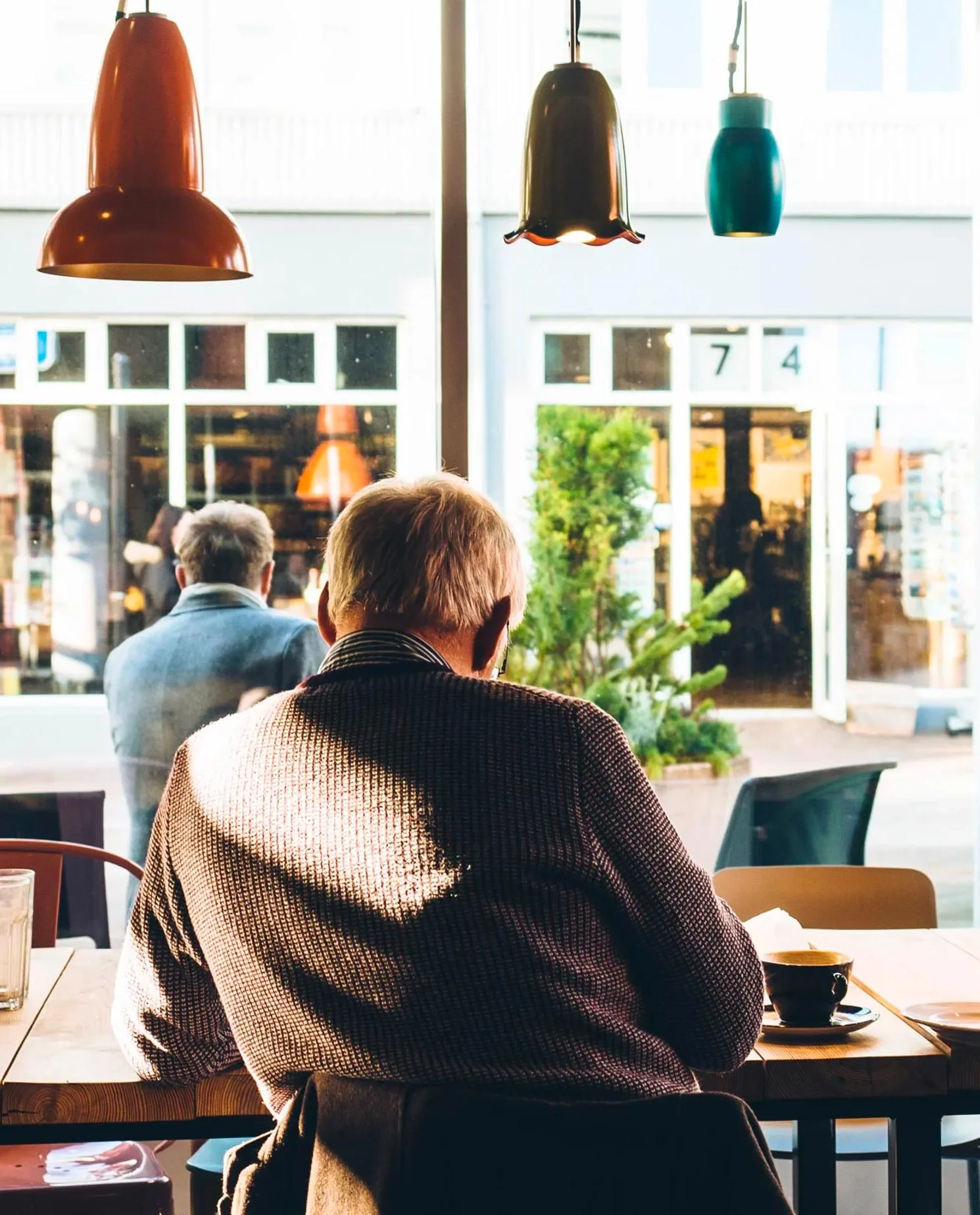 A view of two people sitting in a café, with one person in the foreground facing away, enjoying a coffee and the sunlight filtering through the window.