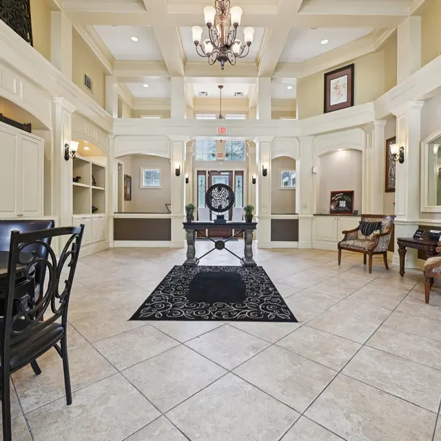 A spacious and elegantly designed lobby featuring high ceilings, a chandelier, and decorative furniture. The floors are tiled, with a central black rug and two seating areas with stylish chairs.