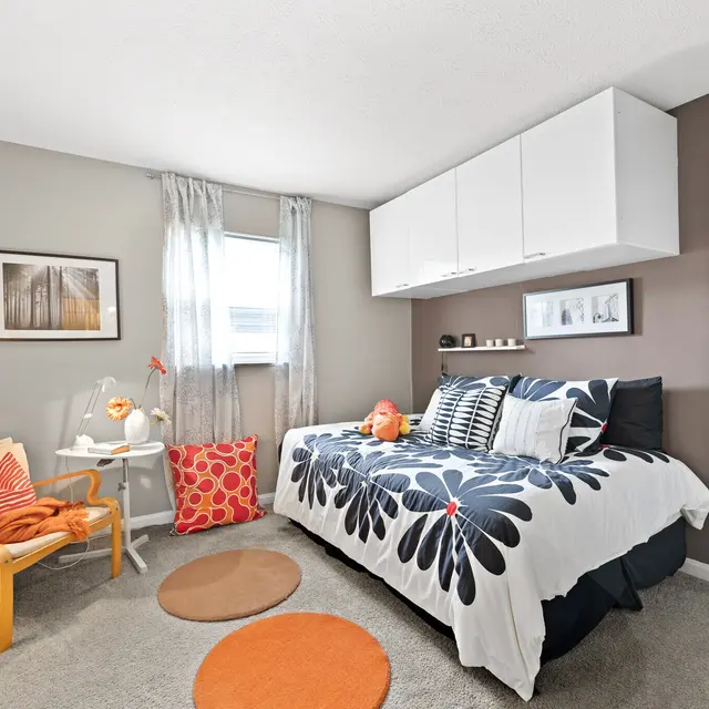 A small, stylish bedroom featuring a double bed with floral bedding, a bright lamp, a wooden chair, and decor elements in warm colors.