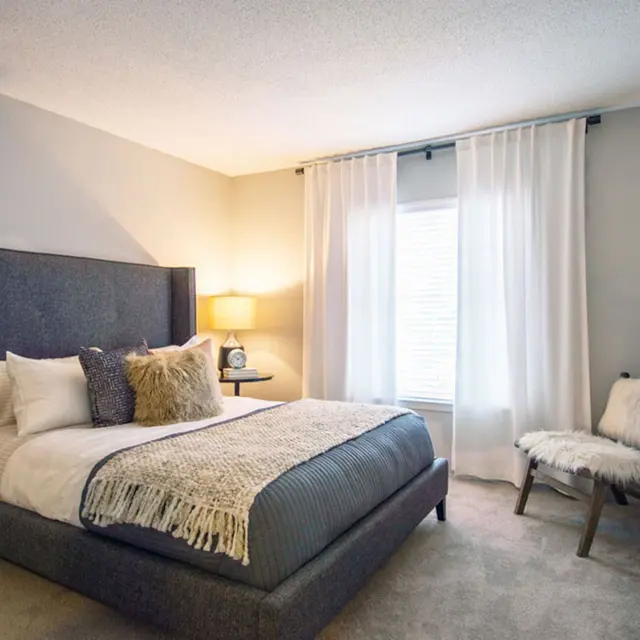 Furnished bedroom with white walls, carpeted flooring and a large window for natural lighting. 