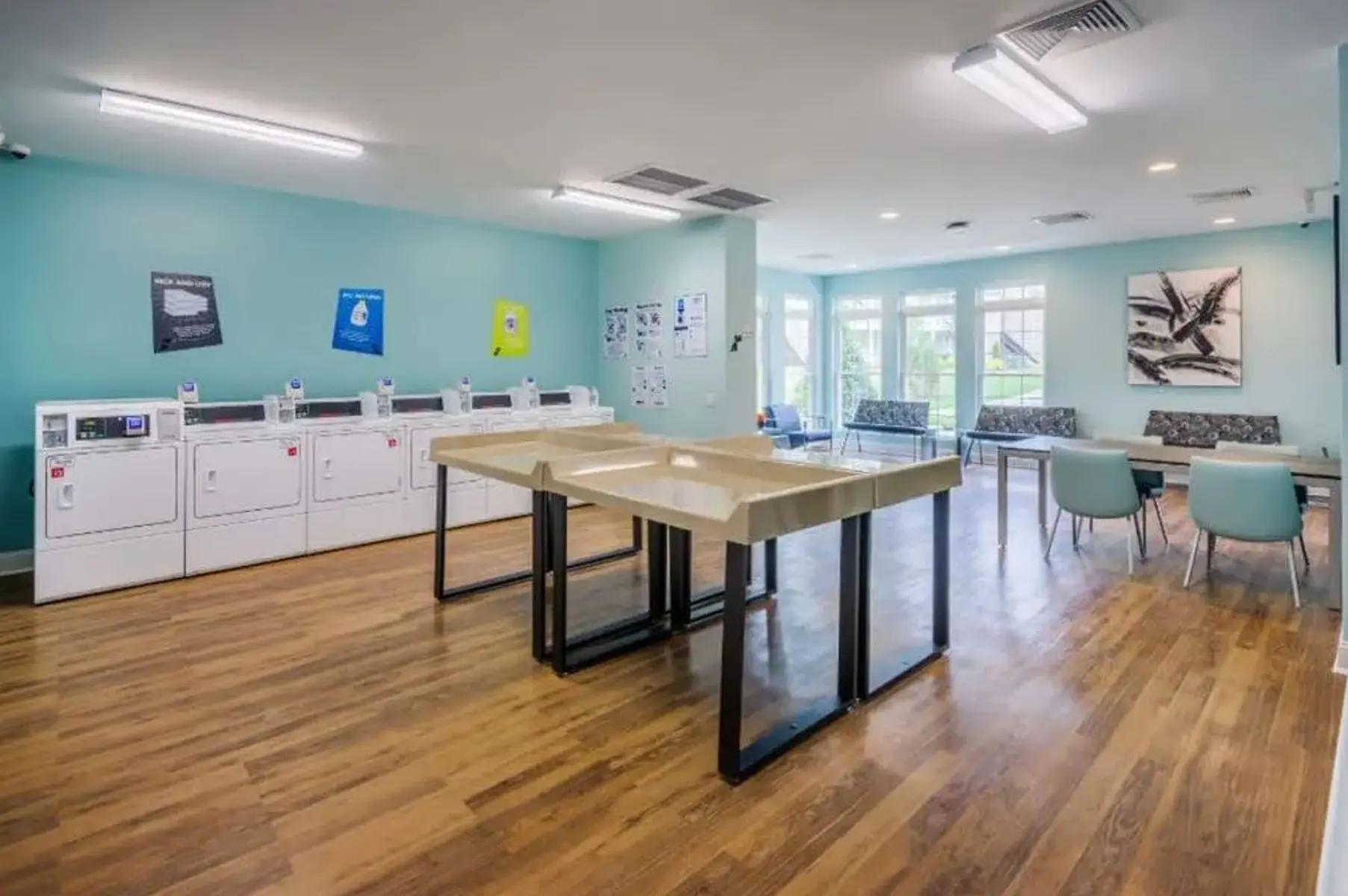 A spacious laundry room with light blue walls and wooden flooring, featuring several washing machines and dryers along one wall. In the center, there is a large folding table, and to the right, there are several tables and chairs for seating. Large windows allow natural light to brighten the space.
