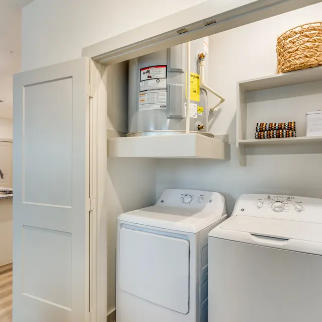 A laundry room featuring a stacked washer and dryer, a water heater mounted above, and a shelf with a woven basket and accessories.
