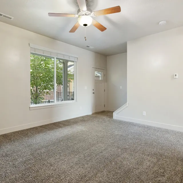 Cozy Living Room Interior A carpeted living room with a ceiling fan and large window allowing natural light. A doorway is visible leading to another room and greenery can be seen outside.