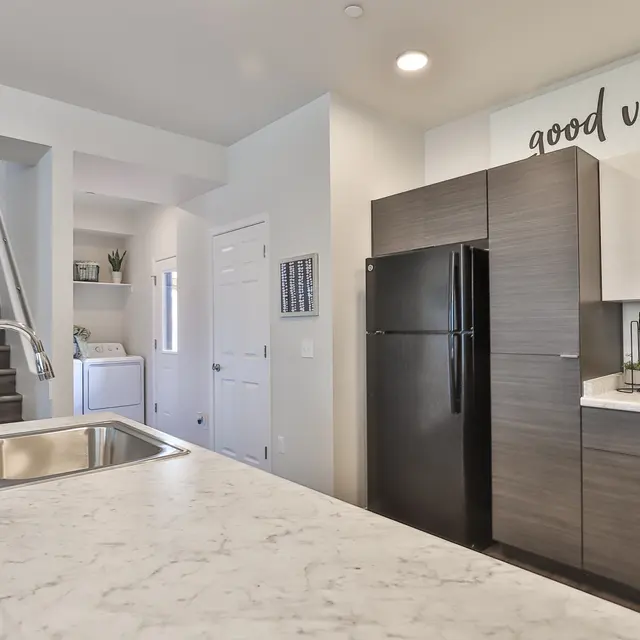 A modern kitchen featuring a marble countertop and dark cabinetry, with a black refrigerator. The walls are white, and there's a motivational phrase 'good vibes' on one of the walls. A staircase is visible in the background, along with some decorative plants.