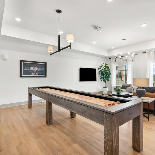 A spacious game room featuring a shuffleboard table, a comfortable seating area with a couch, and large windows with flowing curtains.