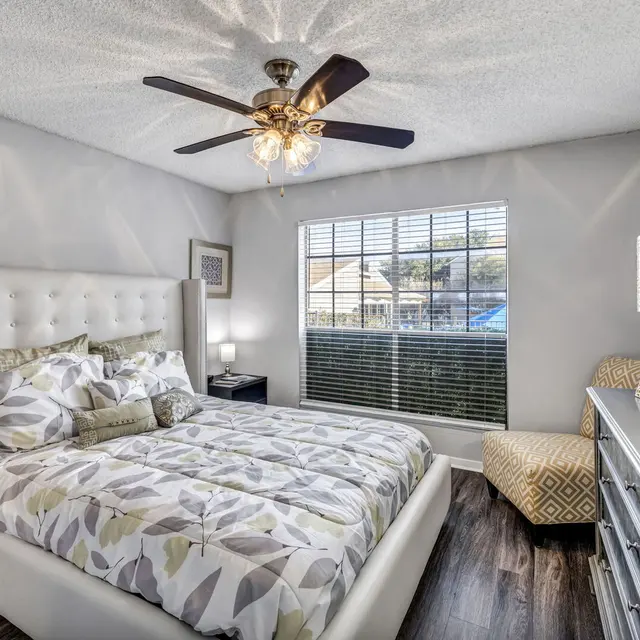 Cozy bedroom featuring a large bed with decorative pillows, a stylish lamp, a fan, and natural light from a window.