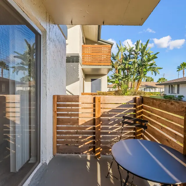 A cozy balcony with a wooden railing, featuring a small black round table and a chair, overlooking palm trees and a residential area.