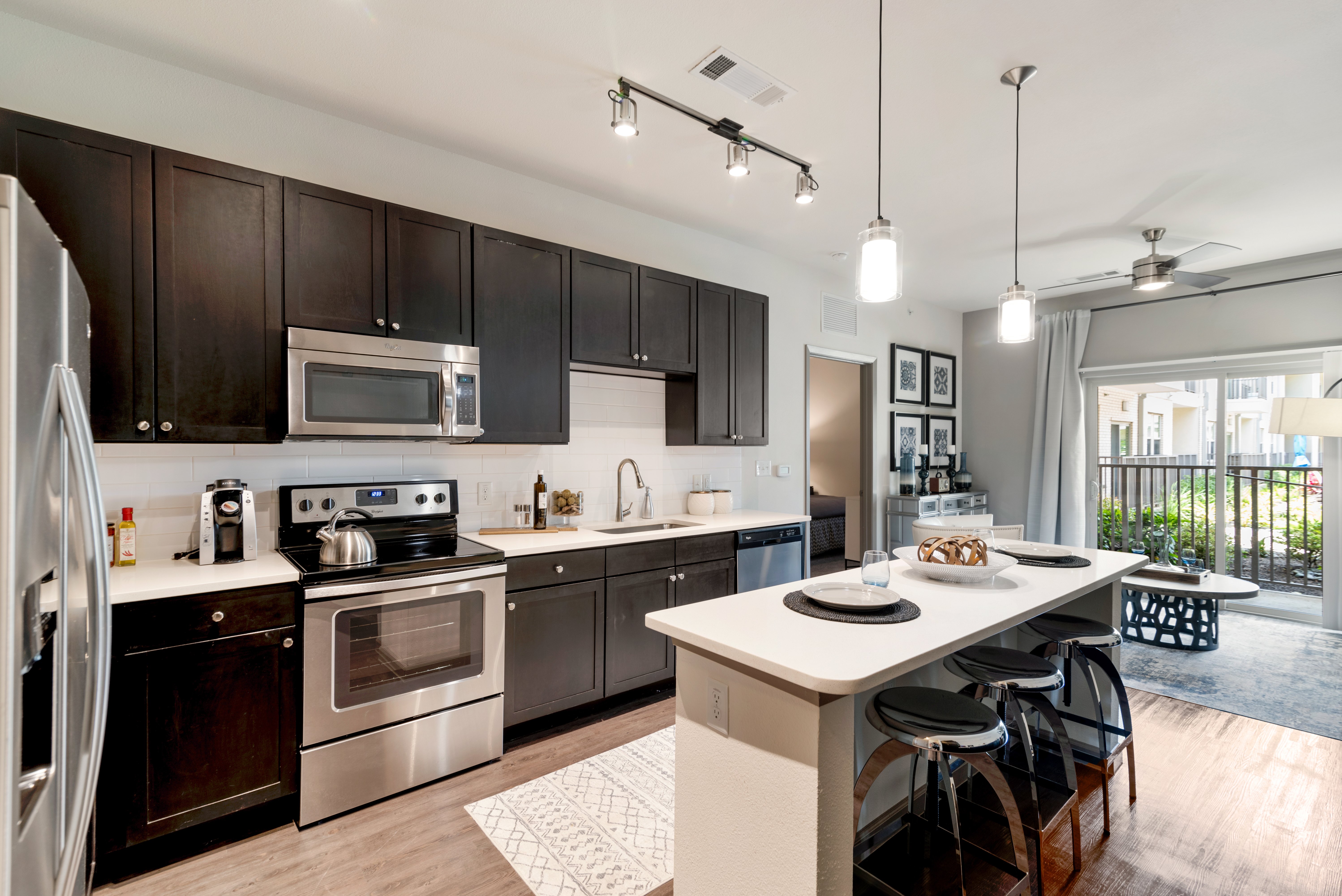 A modern kitchen with dark wooden cabinets, stainless steel appliances, and a countertop with bar stools. A sliding door in the background leads to an outdoor patio area.
