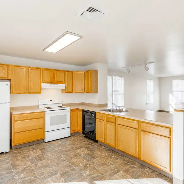 Modern Kitchen Interior A spacious kitchen with wooden cabinets, a white refrigerator, a white stove, and a black dishwasher. The kitchen features a large countertop with a sink, and there is a living area visible in the background with large windows.