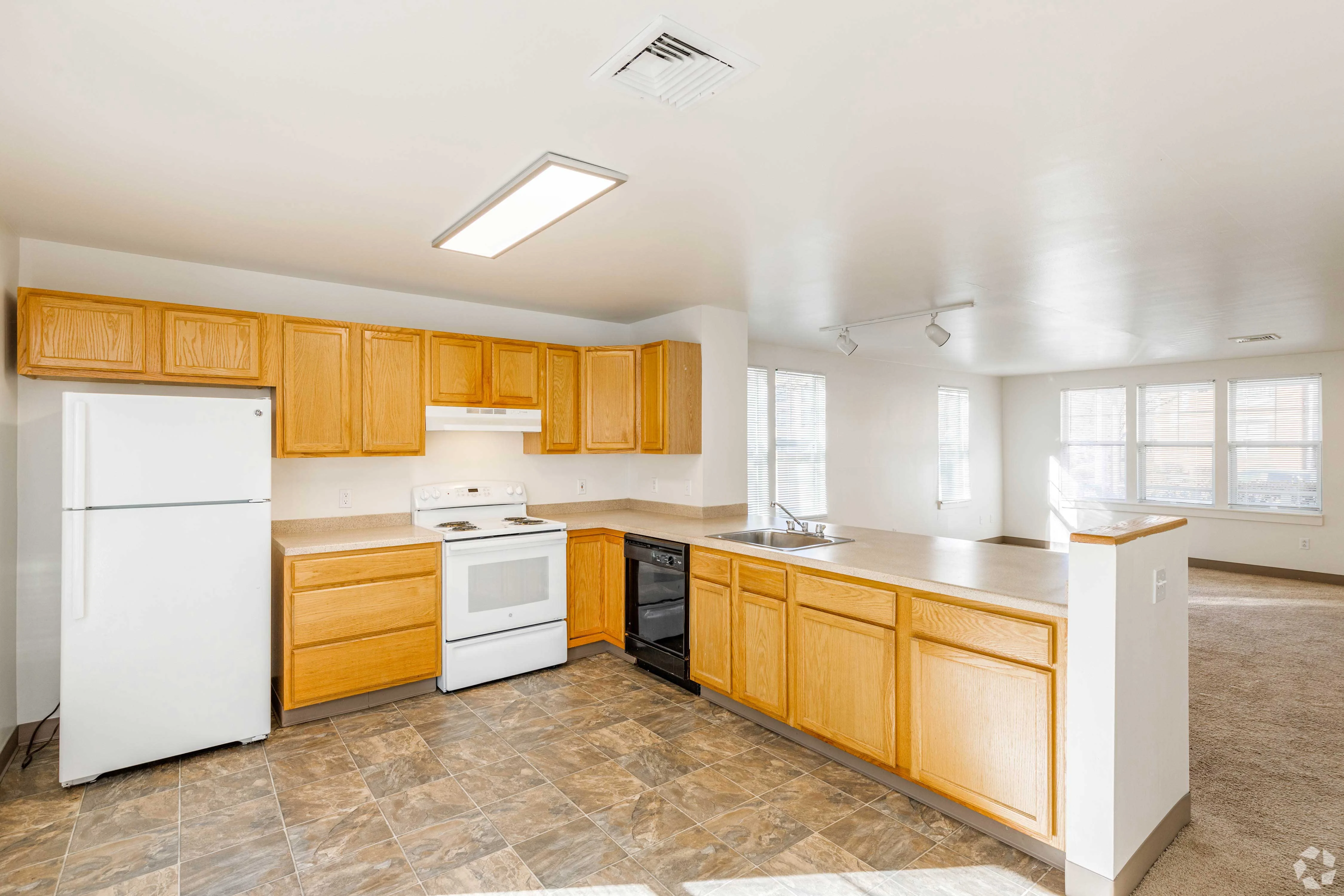 Modern Kitchen Interior A spacious kitchen with wooden cabinets, a white refrigerator, a white stove, and a black dishwasher. The kitchen features a large countertop with a sink, and there is a living area visible in the background with large windows.