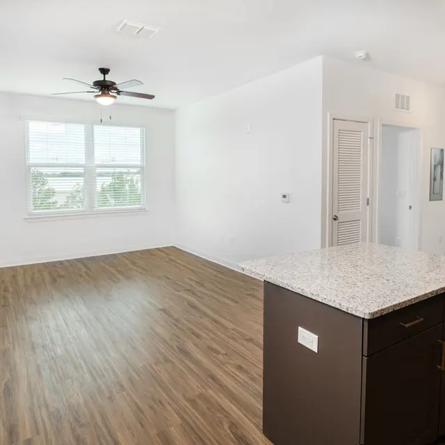 A spacious, modern apartment living area featuring a light-colored wall, a ceiling fan, large windows, and a granite countertop island.