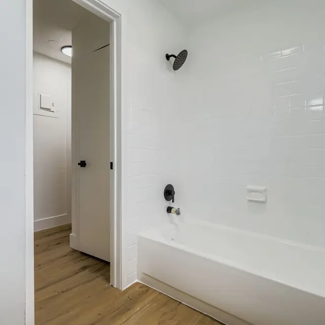 A modern bathroom featuring a bathtub, white tiled walls, and a wooden floor. The door to another room is visible on the left side.