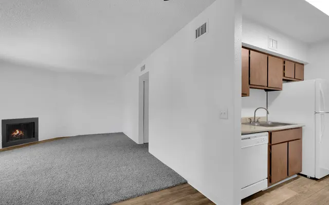 A bright and spacious interior of an apartment featuring a living area with gray carpet, a corner fireplace, and a kitchen area with wooden cabinets, a white refrigerator, and a dishwasher.