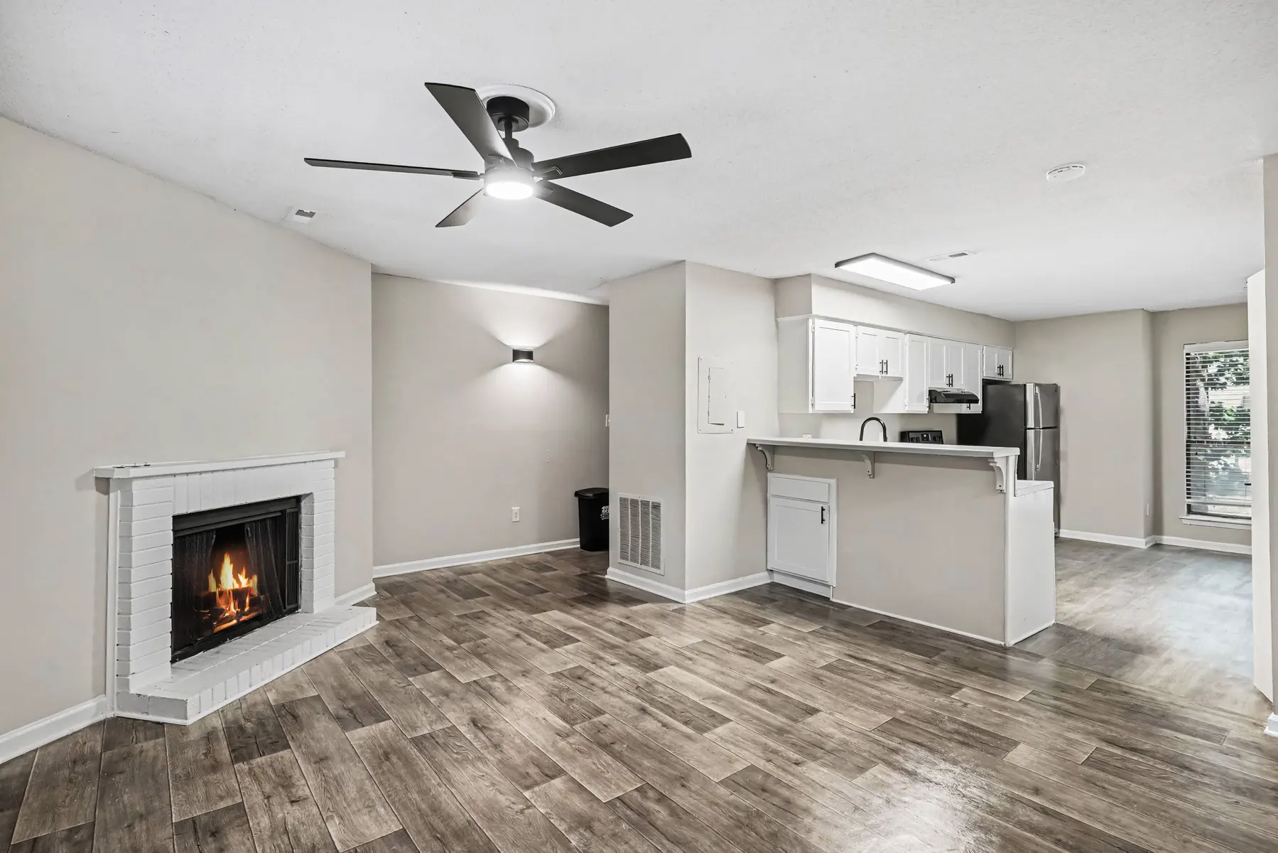 A modern apartment interior featuring a living area with wooden flooring, a fireplace, a ceiling fan, and an open kitchen in the background.