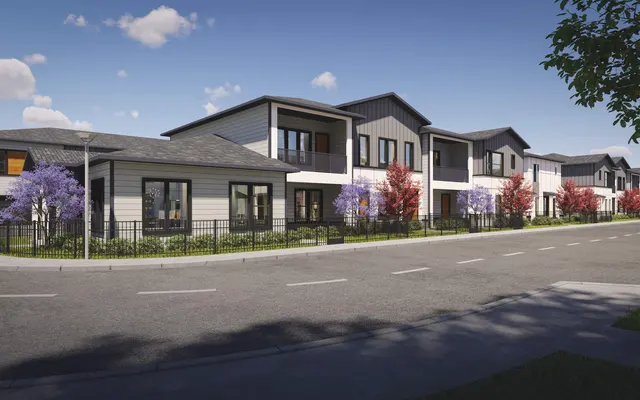 A street view of a modern residential area showcasing several townhouses with contemporary architecture. The buildings are decorated with vibrant foliage in various colors, surrounded by a well-maintained lawn. A clear blue sky with scattered clouds is visible above.