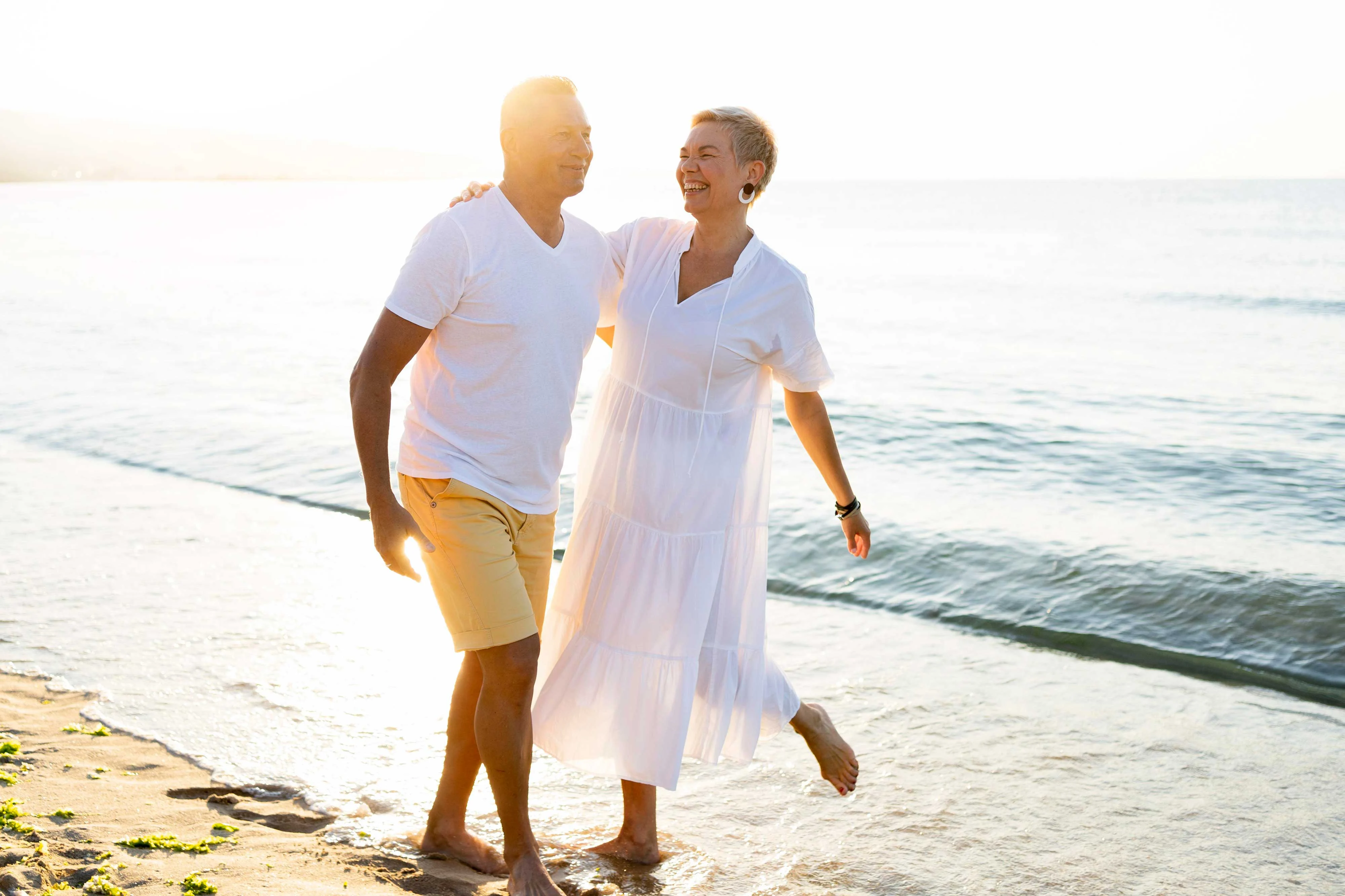 A couple walking barefoot on a beach during sunset. The man is wearing a white shirt and light shorts, while the woman is in a flowing white dress, both smiling and enjoying each other's company.