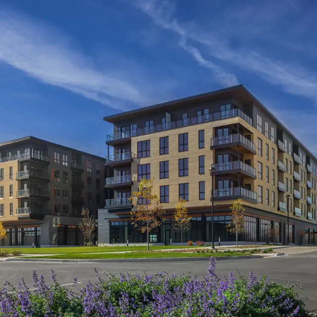 Two modern apartment buildings with balconies, surrounded by green space and blooming plants in the foreground. Blue sky with scattered clouds in the background.