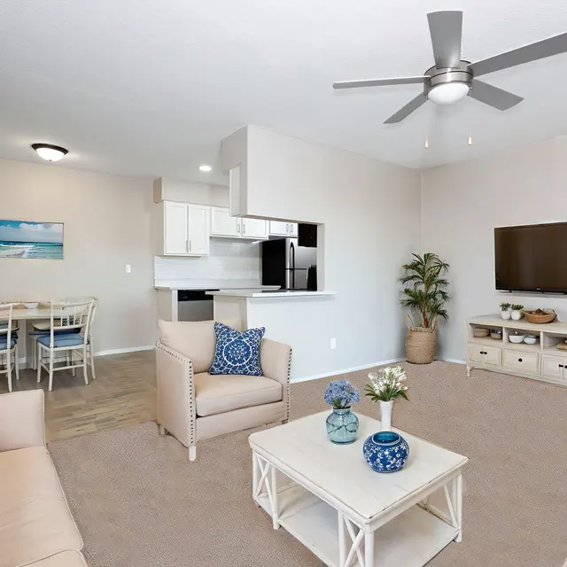 A cozy living room featuring beige seating, a coffee table, a potted plant, and a modern kitchen with white cabinets in the background.