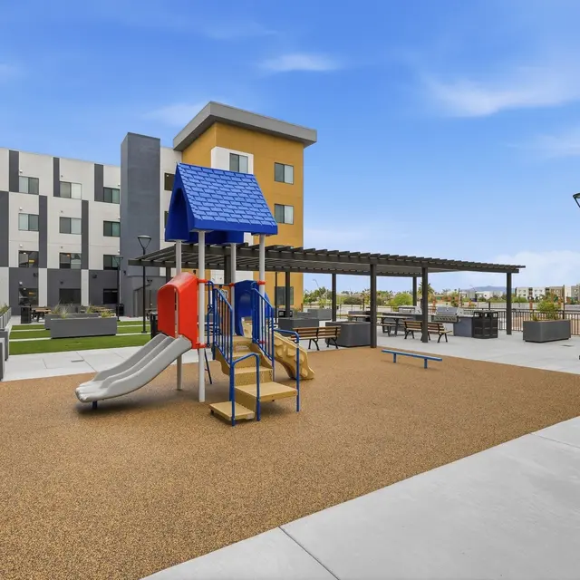Children's Playground in Residential Courtyard View of a playground with slide and climbing structure in a residential courtyard, surrounded by gray and yellow buildings, with seating areas and green space in the background.