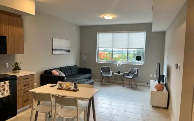 A modern apartment interior featuring a kitchen area with wooden cabinetry, a black couch in the living space, a dining table with two chairs, and a window view of greenery outside.