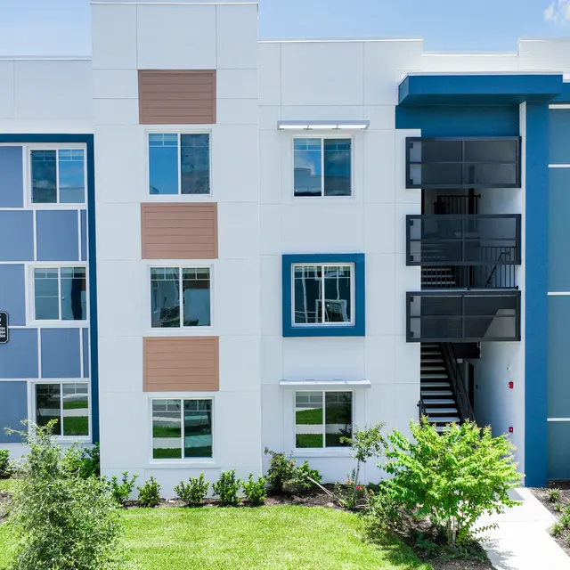 A modern multi-story apartment building with a mix of white and blue exteriors, featuring various window designs and landscaping.