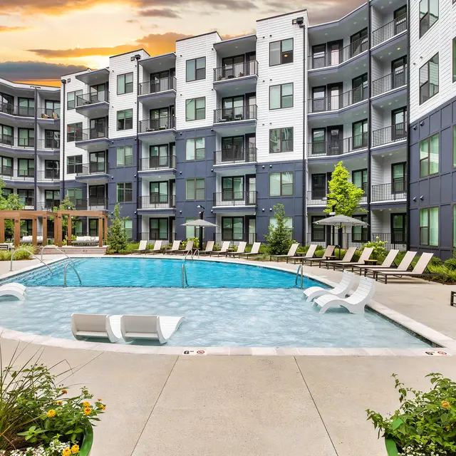 Modern Apartment Pool Area A modern apartment complex courtyard featuring a swimming pool surrounded by lounge chairs and greenery, with an attractive sunset in the background.