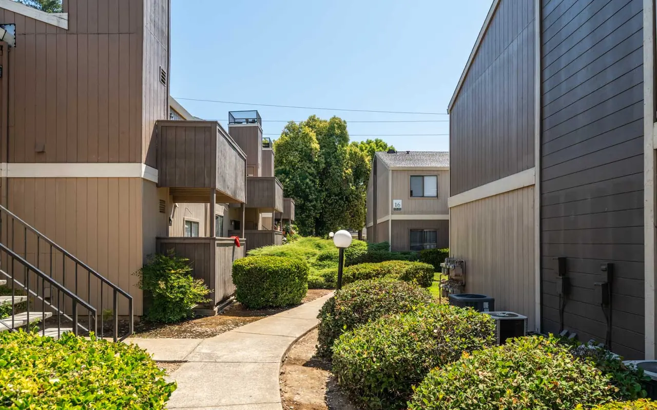 A quiet residential complex featuring a paved pathway surrounded by manicured bushes and two-story buildings under a clear blue sky.