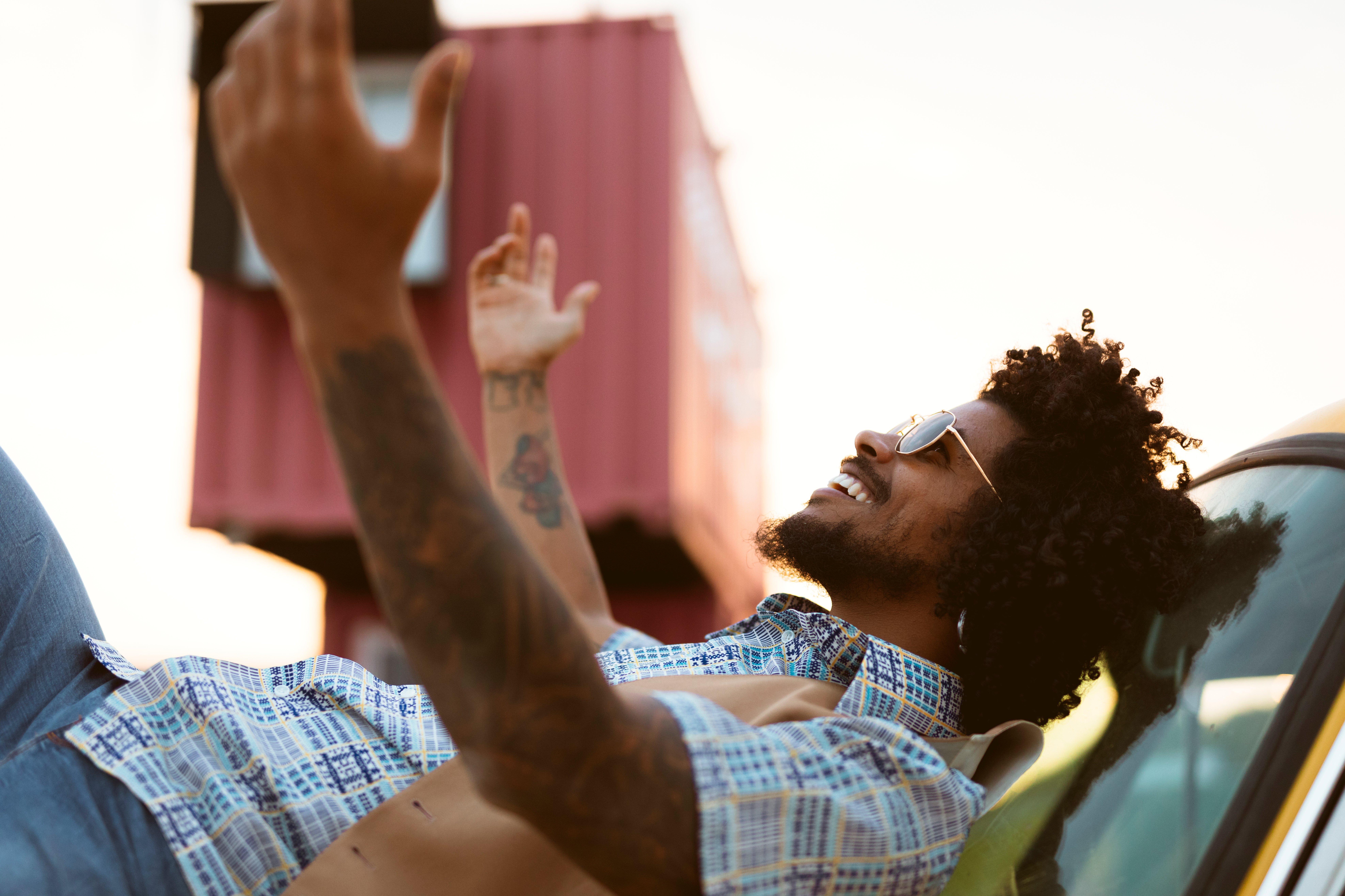 Outdoor Relaxation A man with curly hair wearing sunglasses and a patterned shirt is reclining on a car roof, gesturing joyfully with his arms raised. The background features a container and an outdoor setting during sunset.