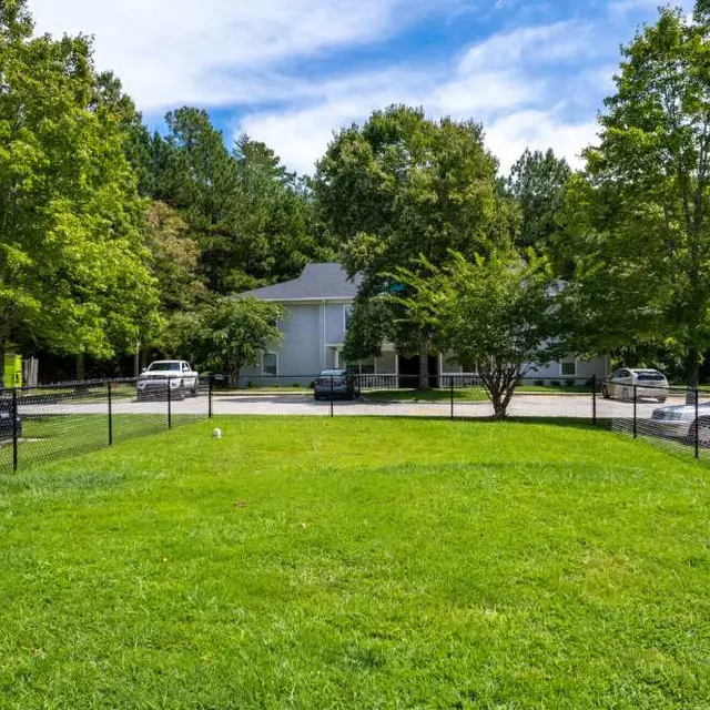 A lush green park area enclosed by a fence, featuring a few cars parked nearby and houses in the background under a blue sky with clouds.